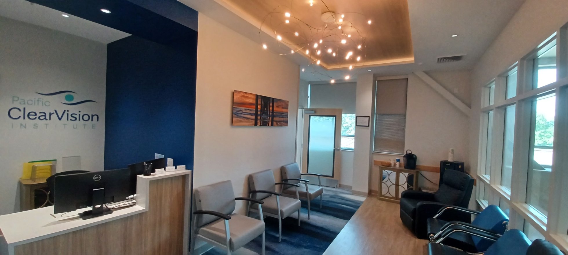 Main Office Waiting Area - Pacific ClearVision Institute Interior view of the PCVI's waiting area in Eugene, OR, featuring modern gray chairs, low wooden tables, a blue-patterned carpet, and a display of eyeglass frames, creating a comfortable and welcoming atmosphere for patients. - Pacific ClearVision Institute